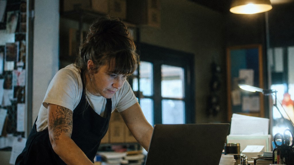 A small-business owner in an apron leaning over a laptop on a cluttered back-office desk late in the day, photographed in Baton Rouge