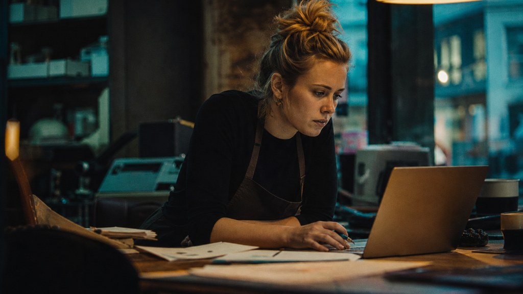 A small-business owner in an apron leaning over a laptop on a cluttered back-office desk late in the day, photographed in Belfast