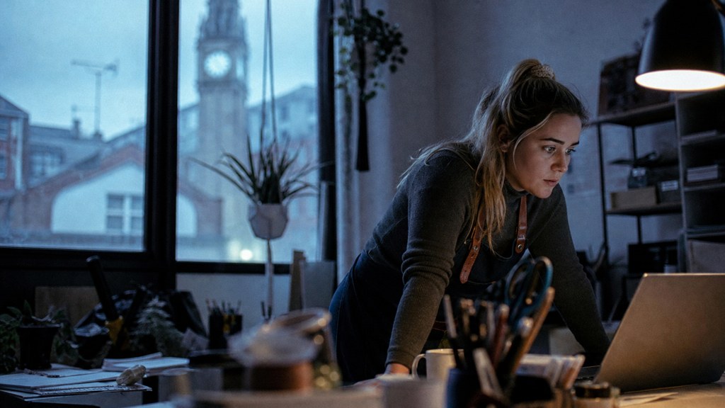 A small-business owner in an apron leaning over a laptop on a cluttered back-office desk late in the day, photographed in Belfast