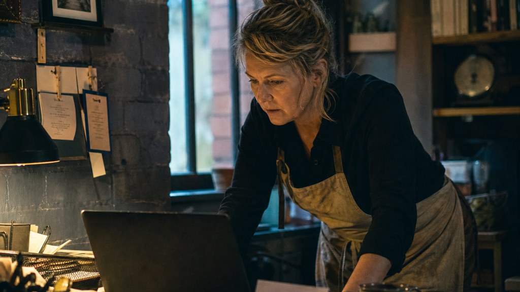 A small-business owner in an apron leaning over a laptop on a cluttered back-office desk late in the day, photographed in Belfast
