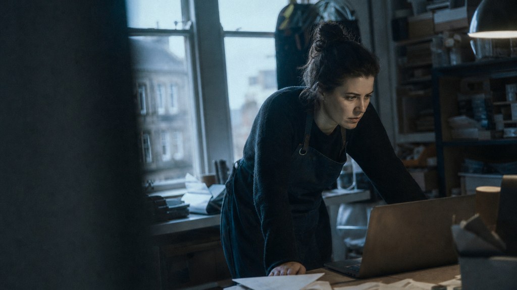 A small-business owner in an apron leaning over a laptop on a cluttered back-office desk late in the day, photographed in Belfast