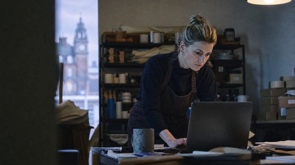 A small-business owner in an apron leaning over a laptop on a cluttered back-office desk late in the day, photographed in Belfast