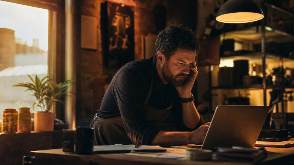 A small-business owner in an apron leaning over a laptop on a cluttered back-office desk late in the day, photographed in Birmingham