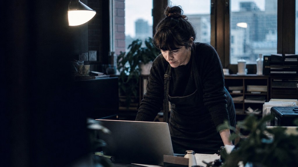 A small-business owner in an apron leaning over a laptop on a cluttered back-office desk late in the day, photographed in Birmingham