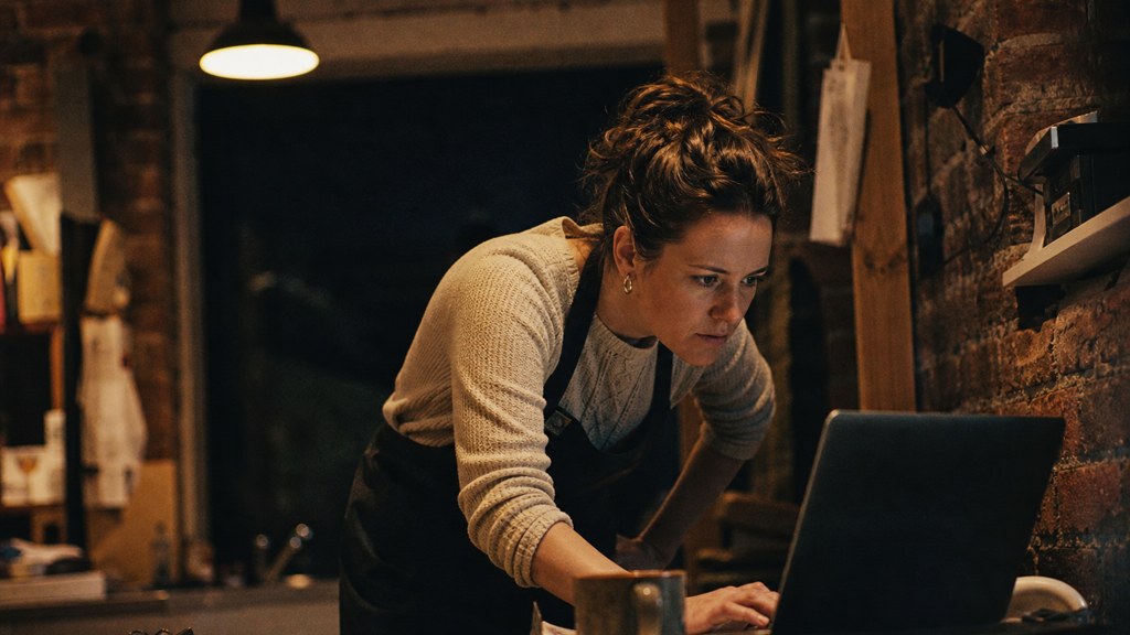 A small-business owner in an apron leaning over a laptop on a cluttered back-office desk late in the day, photographed in Birmingham