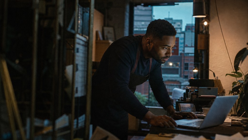 A small-business owner in an apron leaning over a laptop on a cluttered back-office desk late in the day, photographed in Birmingham