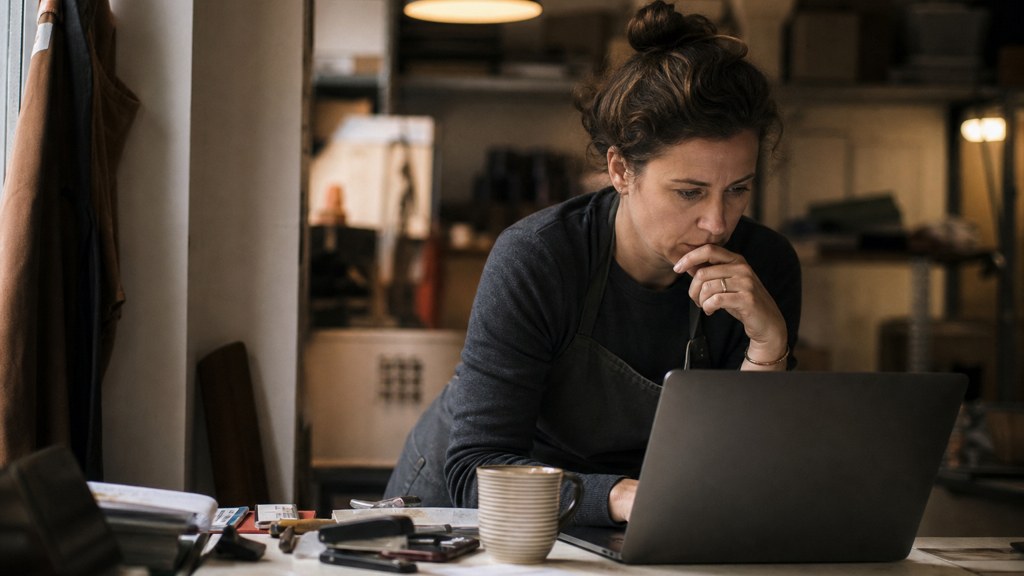 A small-business owner in an apron leaning over a laptop on a cluttered back-office desk late in the day, photographed in Birmingham
