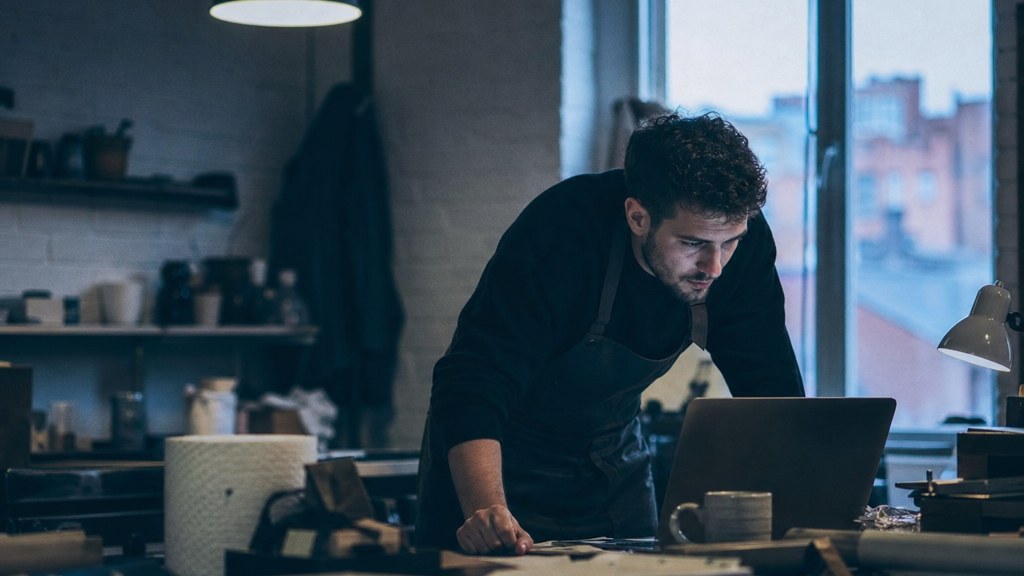 A small-business owner in an apron leaning over a laptop on a cluttered back-office desk late in the day, photographed in Birmingham