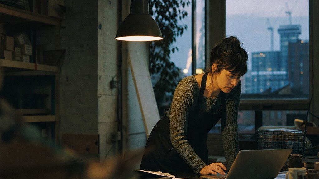A small-business owner in an apron leaning over a laptop on a cluttered back-office desk late in the day, photographed in Birmingham