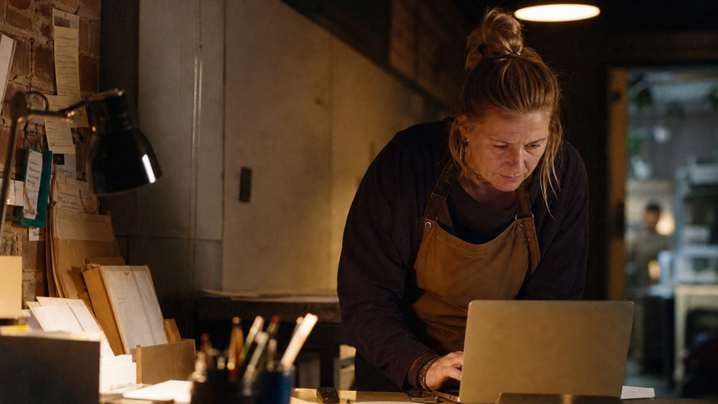 A small-business owner in an apron leaning over a laptop on a cluttered back-office desk late in the day, photographed in Birmingham
