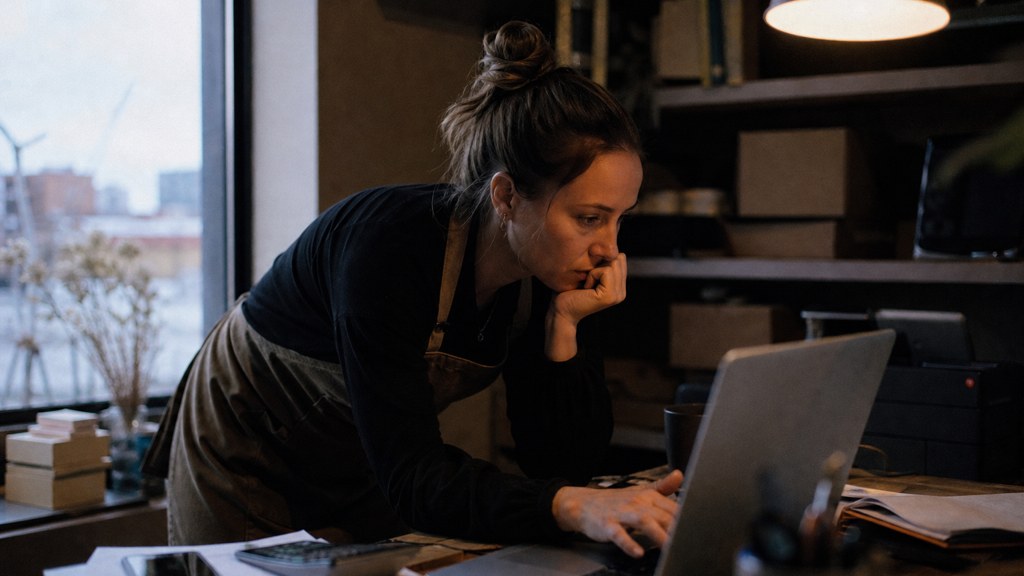 A small-business owner in an apron leaning over a laptop on a cluttered back-office desk late in the day, photographed in Birmingham