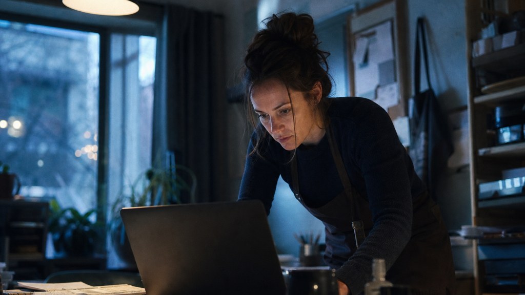 A small-business owner in an apron leaning over a laptop on a cluttered back-office desk late in the day, photographed in Boise