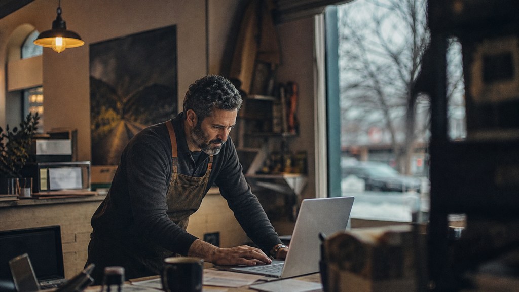 A small-business owner in an apron leaning over a laptop on a cluttered back-office desk late in the day, photographed in Boise