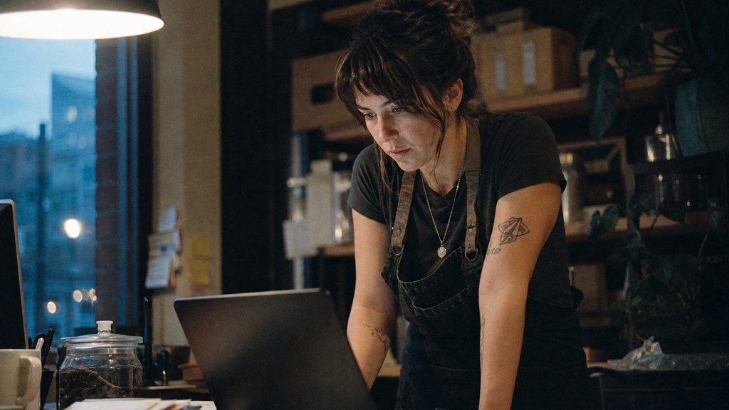 A small-business owner in an apron leaning over a laptop on a cluttered back-office desk late in the day, photographed in Boise