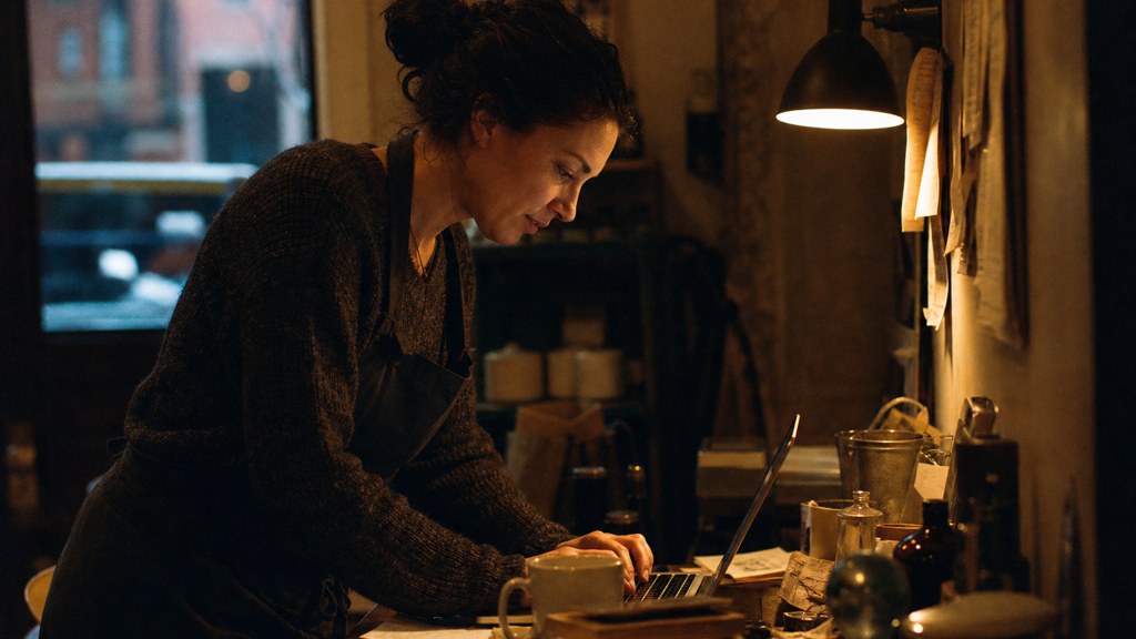 A small-business owner in an apron leaning over a laptop on a cluttered back-office desk late in the day, photographed in Boston