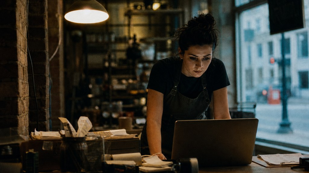 A small-business owner in an apron leaning over a laptop on a cluttered back-office desk late in the day, photographed in Boston