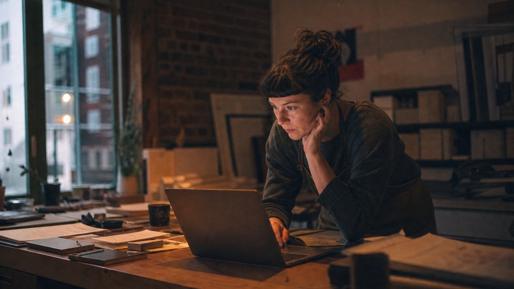 A small-business owner in an apron leaning over a laptop on a cluttered back-office desk late in the day, photographed in Boston