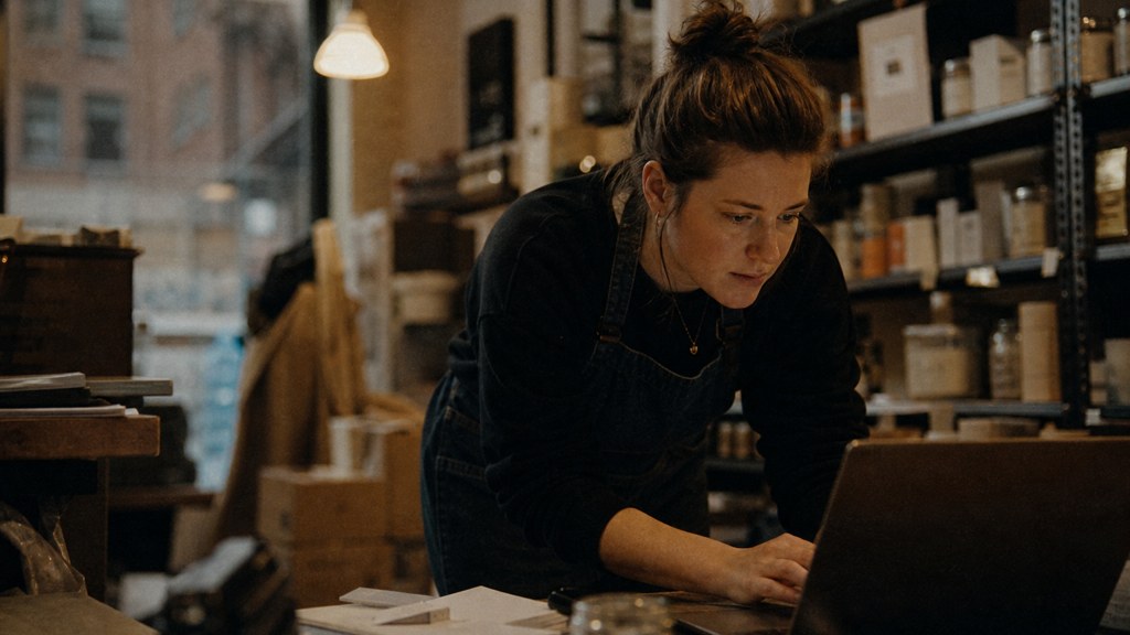 A small-business owner in an apron leaning over a laptop on a cluttered back-office desk late in the day, photographed in Boston