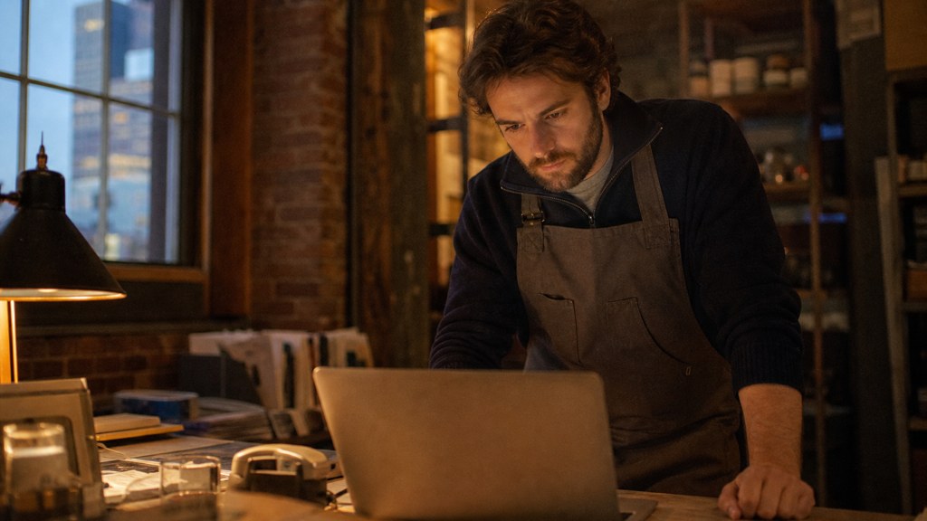 A small-business owner in an apron leaning over a laptop on a cluttered back-office desk late in the day, photographed in Boston