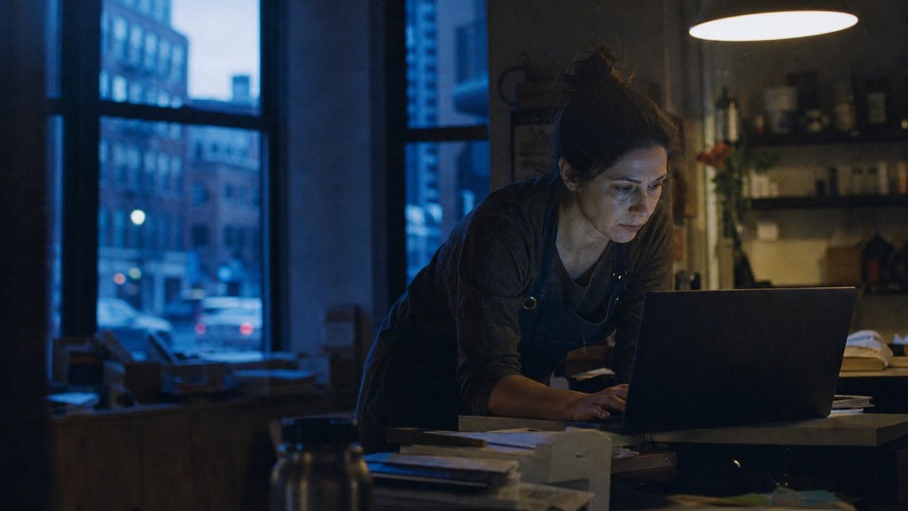 A small-business owner in an apron leaning over a laptop on a cluttered back-office desk late in the day, photographed in Boston