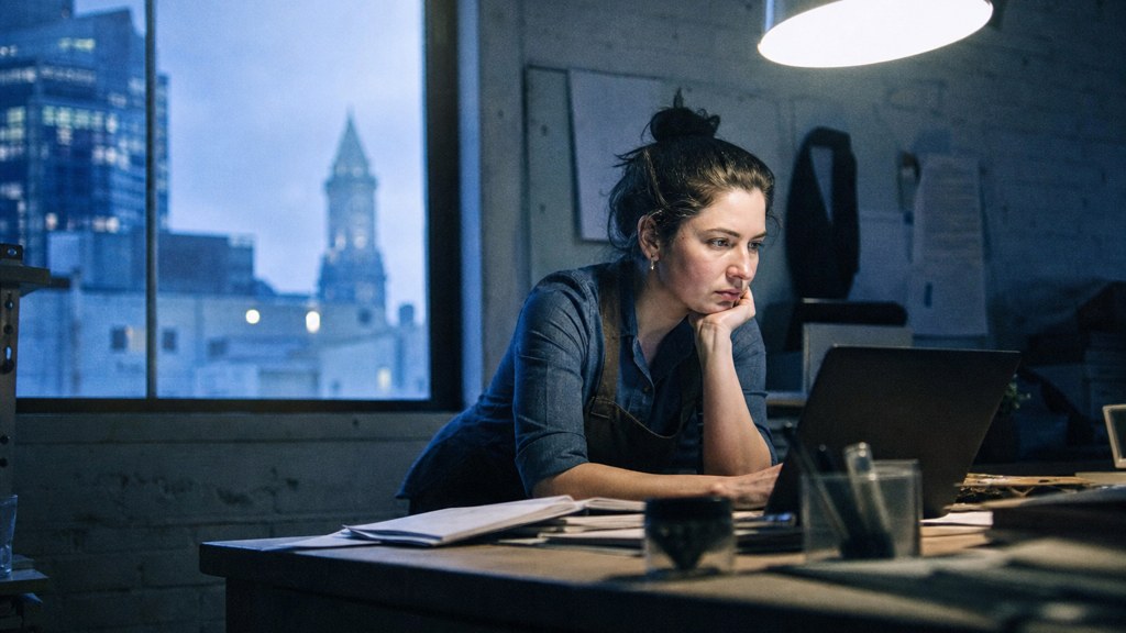 A small-business owner in an apron leaning over a laptop on a cluttered back-office desk late in the day, photographed in Boston