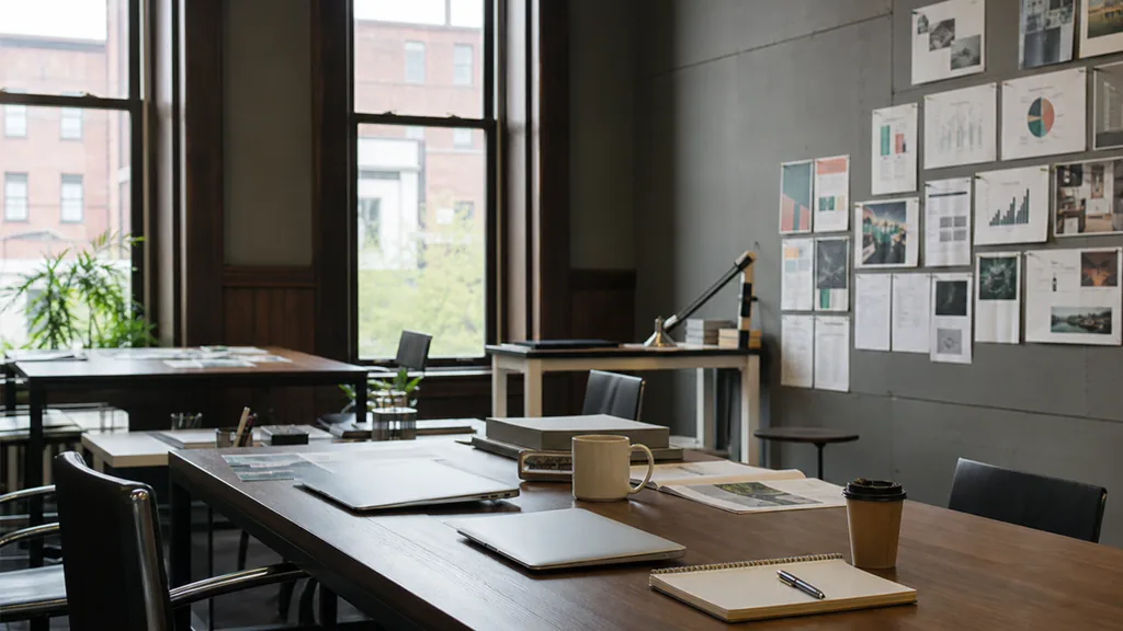 Editorial photograph of an empty modern marketing studio in a Hartford office with dark walnut, tall sash windows and cool New England daylight, a widescreen monitor showing a blank reporting view, a keyboard and a mug on a wooden desk, no people and no signage