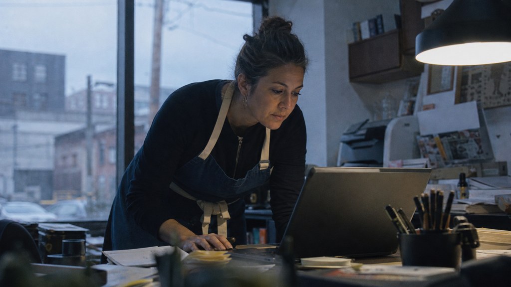A small-business owner in an apron leaning over a laptop on a cluttered back-office desk late in the day, photographed in Bridgeport