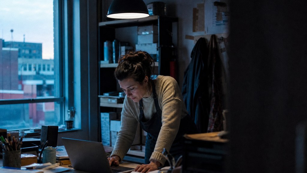 A small-business owner in an apron leaning over a laptop on a cluttered back-office desk late in the day, photographed in Bridgeport