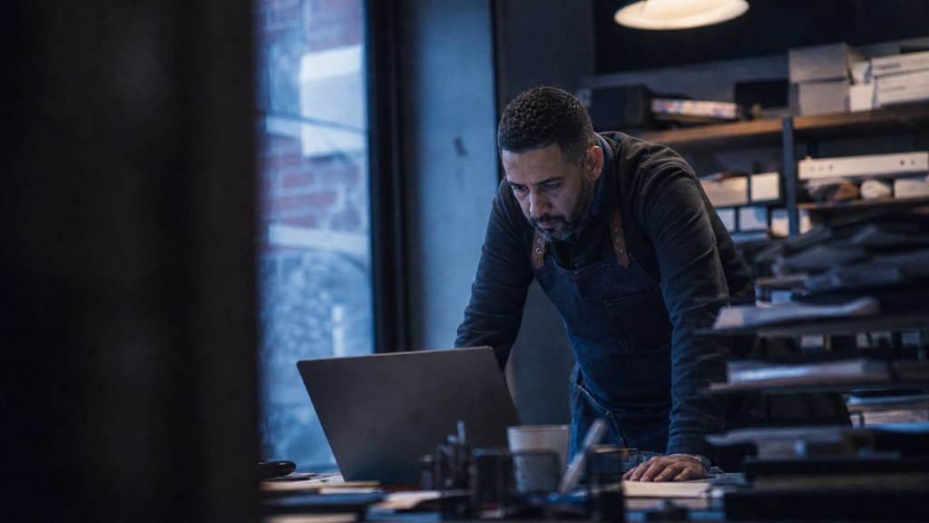 A small-business owner in an apron leaning over a laptop on a cluttered back-office desk late in the day, photographed in Bridgeport