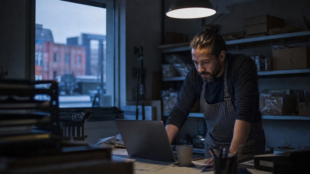 A small-business owner in an apron leaning over a laptop on a cluttered back-office desk late in the day, photographed in Bridgeport