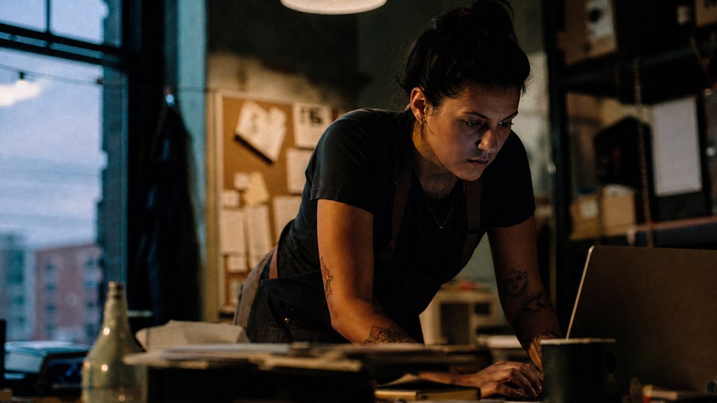 A small-business owner in an apron leaning over a laptop on a cluttered back-office desk late in the day, photographed in Bridgeport