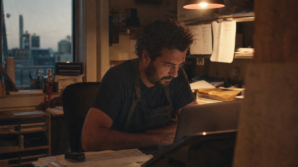 A small-business owner in an apron leaning over a laptop on a cluttered back-office desk late in the day, photographed in Bridgeport