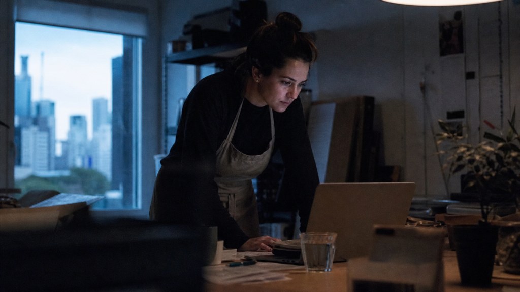 A small-business owner in an apron leaning over a laptop on a cluttered back-office desk late in the day, photographed in Brisbane
