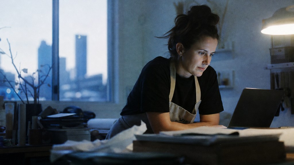 A small-business owner in an apron leaning over a laptop on a cluttered back-office desk late in the day, photographed in Brisbane