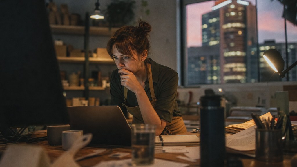 A small-business owner in an apron leaning over a laptop on a cluttered back-office desk late in the day, photographed in Brisbane