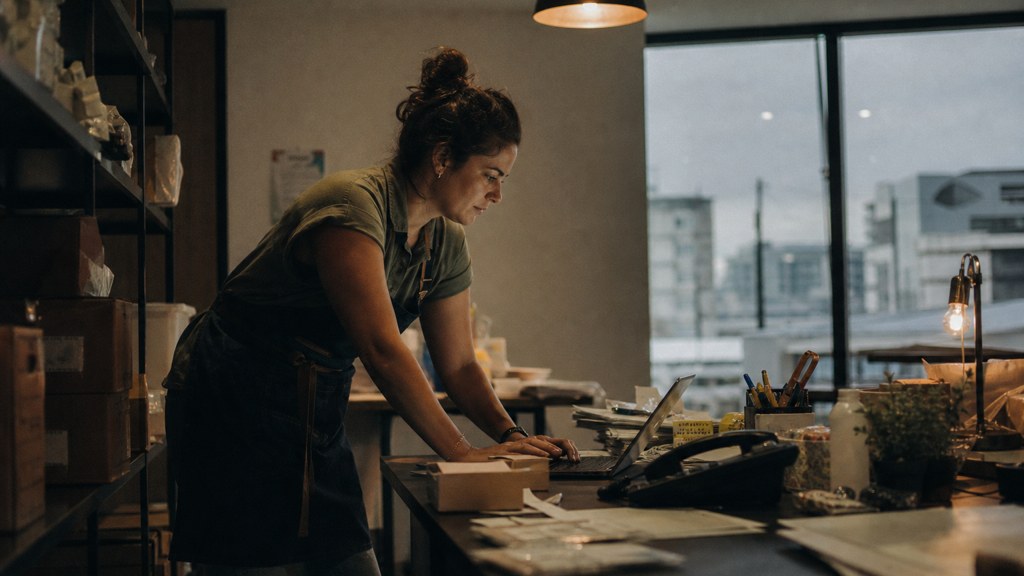 A small-business owner in an apron leaning over a laptop on a cluttered back-office desk late in the day, photographed in Brisbane