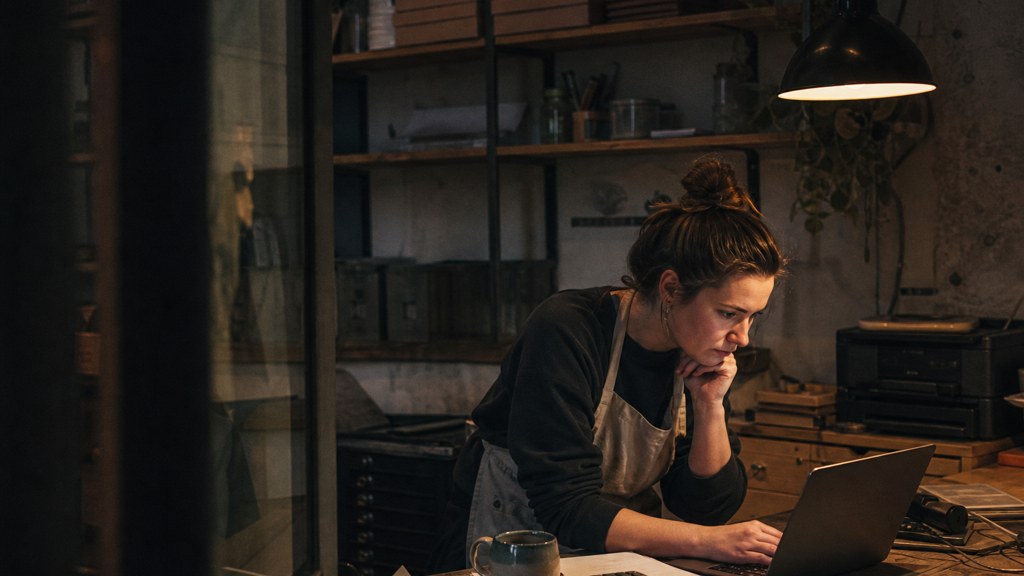 A small-business owner in an apron leaning over a laptop on a cluttered back-office desk late in the day, photographed in Bristol