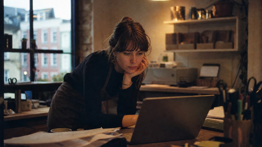 A small-business owner in an apron leaning over a laptop on a cluttered back-office desk late in the day, photographed in Bristol