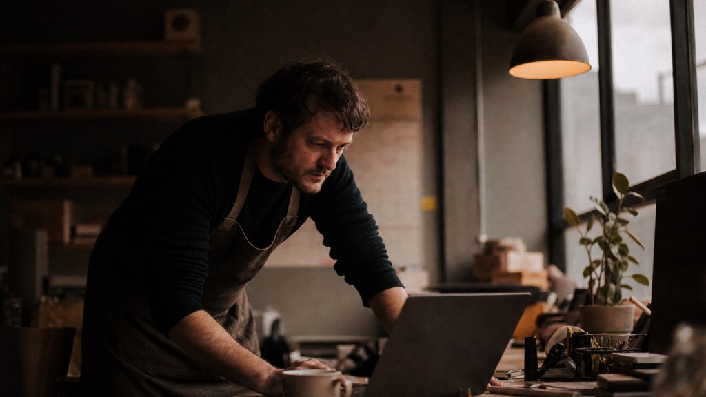 A small-business owner in an apron leaning over a laptop on a cluttered back-office desk late in the day, photographed in Bristol