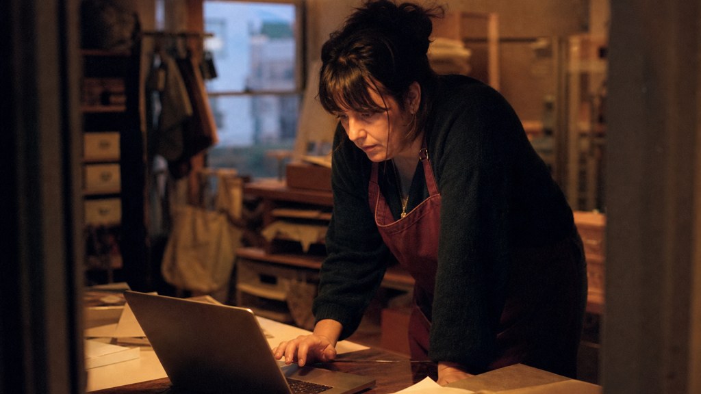 A small-business owner in an apron leaning over a laptop on a cluttered back-office desk late in the day, photographed in Bristol
