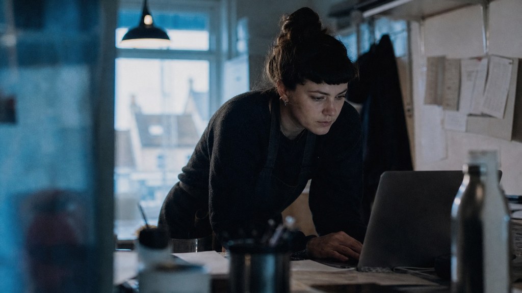 A small-business owner in an apron leaning over a laptop on a cluttered back-office desk late in the day, photographed in Bristol