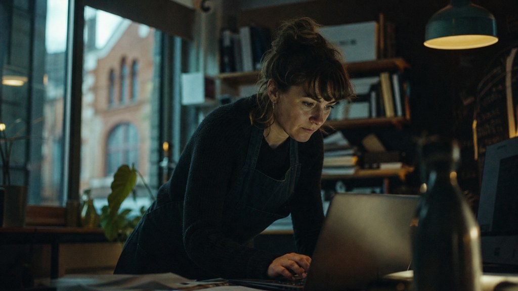 A small-business owner in an apron leaning over a laptop on a cluttered back-office desk late in the day, photographed in Bristol