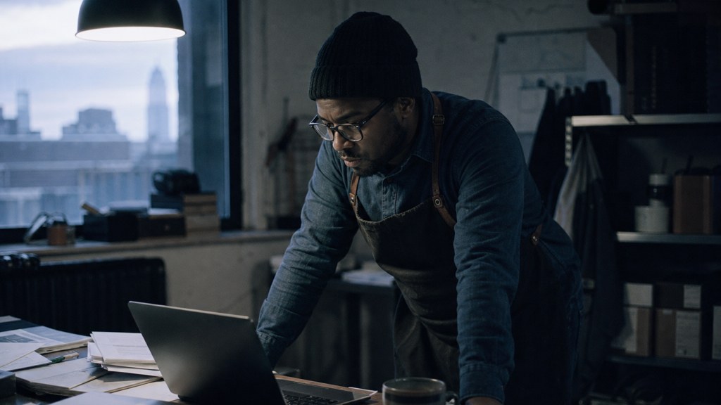 A small-business owner in an apron leaning over a laptop on a cluttered back-office desk late in the day, photographed in Buffalo