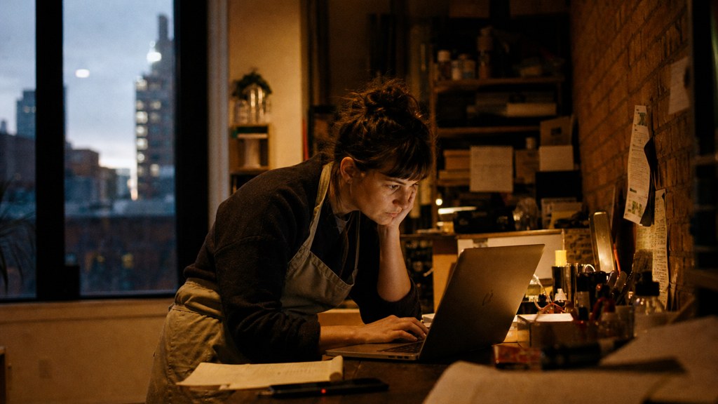 A small-business owner in an apron leaning over a laptop on a cluttered back-office desk late in the day, photographed in Buffalo