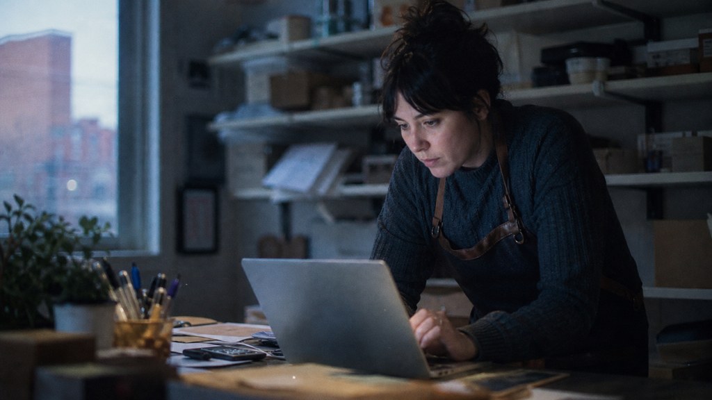 A small-business owner in an apron leaning over a laptop on a cluttered back-office desk late in the day, photographed in Buffalo