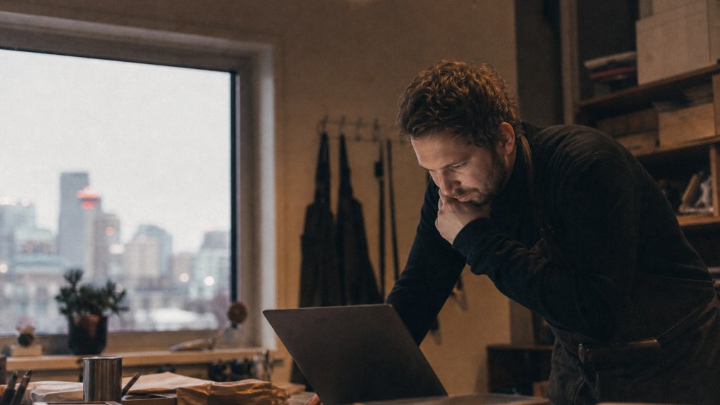 A small-business owner in an apron leaning over a laptop on a cluttered back-office desk late in the day, photographed in Calgary