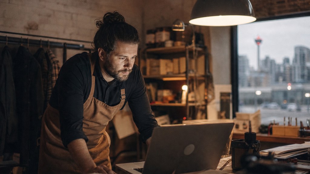 A small-business owner in an apron leaning over a laptop on a cluttered back-office desk late in the day, photographed in Calgary