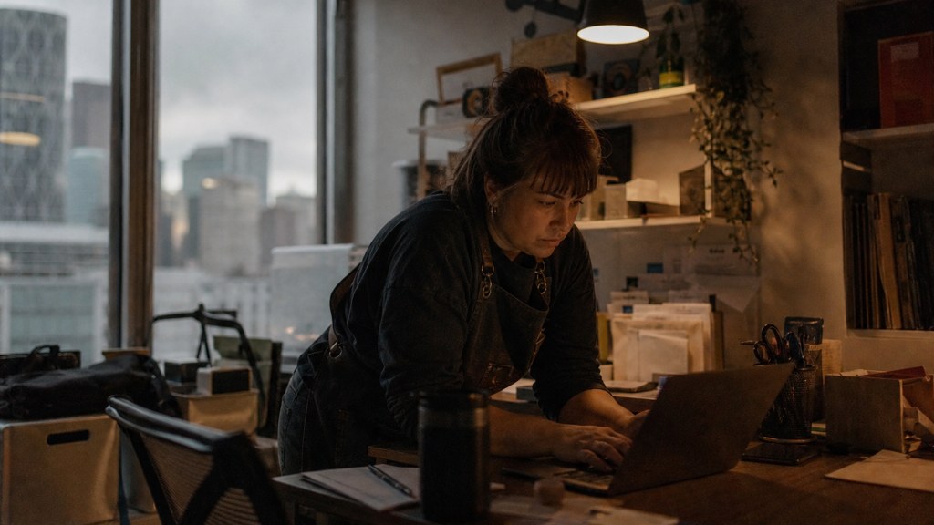 A small-business owner in an apron leaning over a laptop on a cluttered back-office desk late in the day, photographed in Calgary
