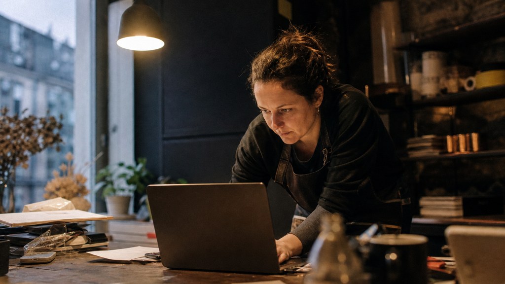 A small-business owner in an apron leaning over a laptop on a cluttered back-office desk late in the day, photographed in Cardiff