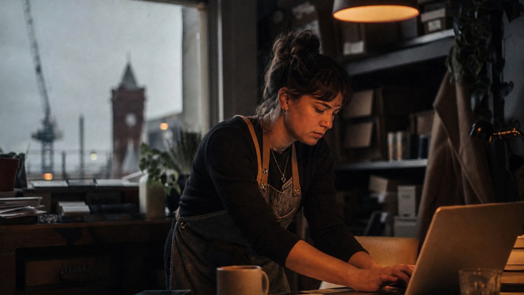 A small-business owner in an apron leaning over a laptop on a cluttered back-office desk late in the day, photographed in Cardiff